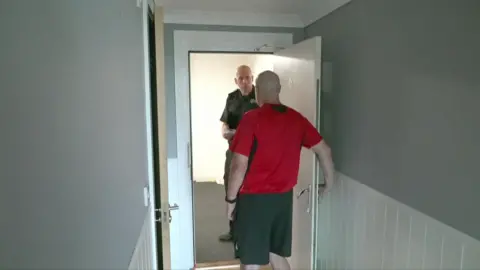 Dave Hemings, who had the camera back, was wearing shorts and an SSA shirt. Ng standing at the door, which he has answered, is the format of sliding and saving the Postage Service wearing long green pants and the same shirt. He had in his hand a copy of the prohibition notice.