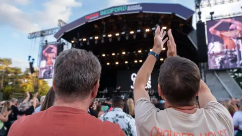 Cuffe & Taylor A close-up of two people in a field, at a concert, one has his arms up, a stage is in front of them, with an act performing on it, other people are around them. 