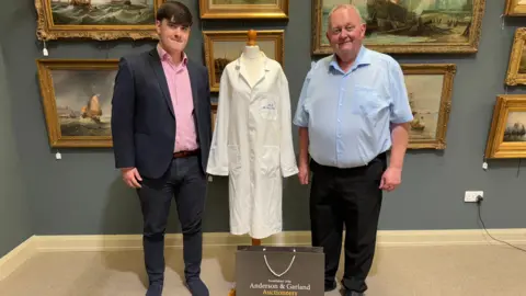 Graham Greeves is standing to the right of the white lab coat which is on a stand. He is wearing a pink shirt and black trousers. There is a man on the right wearing a light blue shirt.