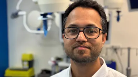 An Asian man with short dark hair and a short grey beard. He is standing in a medical setting and is wearing a white shirt as well as black glasses. He is smiling into the camera.