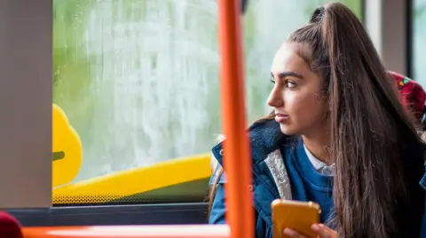 A teenage girl looks out of a bus window. She has long brown hair. She is wearing a blue jumper, blue coat and is holding a smartphone in an orange coloured case.