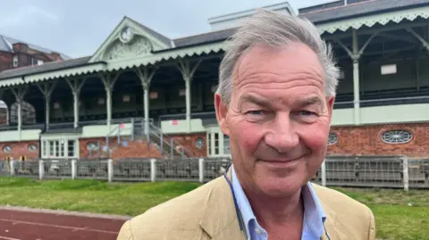 Rupert Lowe standing in front of the Wellesley Football Stand in Great Yarmouth. He has grey hair, wears a gold-coloured suit jacket and blue shirt, and is looking at the camera. The football stand looks Victorian, with a sage green trim to it.