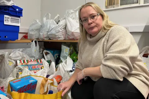 Kevin Shoesmith/BBC Samantha Palmer, a food bank co-ordinator, wearing a cream-coloured pullover and black trousers, crouches down next to dozens of carrier bags full of donated food