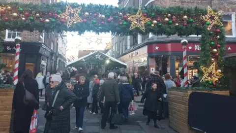 BBC A shot of the Christmas Market on Parliament Square in York, with a large fir tree archway, decorated with gold, green and red baubles and illuminated stars. A mixed crowd of people can be seen walking beneath it.
