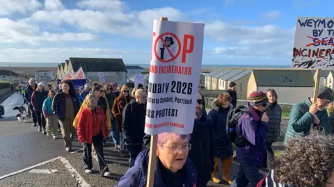 A line of people walking on the coast, two at the front holding banners. 