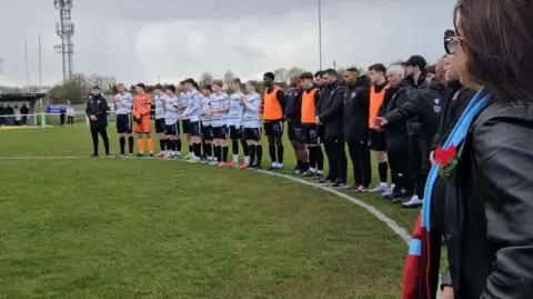 Ashville FC players form a semi-circle on an outdoor football pitch and bow their heads in respect