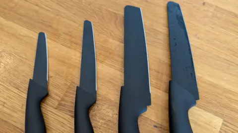 Four rounded-tip charcoal grey kitchen knives of varying size laid out on a wooden table. 