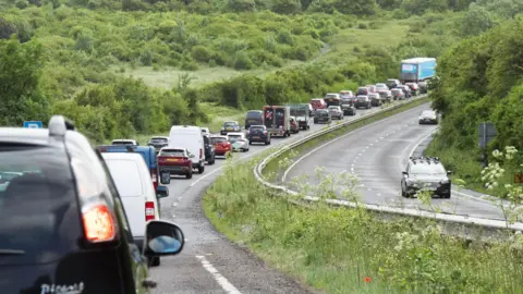 Long queues of cars on one carriageway. The other carriageway closed with surrounding hills. 