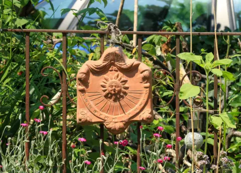 Doris Enders  A clay sundial on metal railings. Wildflowers and greenery are behind the railings