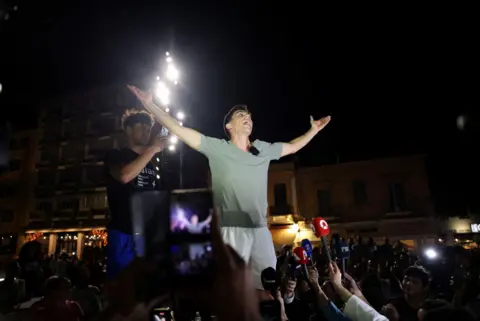 YouTuber Fidias Panayuotou standing in front of some microphones with his arms raised in the air at a rally