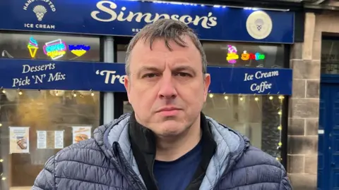 A man with short grey hair wearing a parka jacket standing in front of a shop with blue signage.