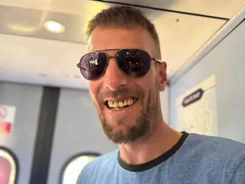 A head and shoulders photograph of passenger David Naden. He is standing on the train and is wearing a blue t-shirt and sunglasses. He has short, dark hair and a short beard.