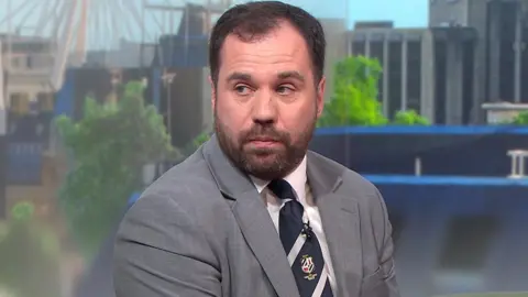 Sean Woodcock has short brown hair and a beard. He's wearing a grey blazer over a white shirt with a blue tie. There is a television studio backdrop behind him.