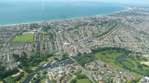 Aerial view over Christchurch, looking towards Bournemouth, Poole and Poole Harbour. The three towns are linked by a long curved sandy beach which runs along the top of the picture with the sea merging with the sky on a misty horizon. Directly below the photographer is the meandering river Avon.