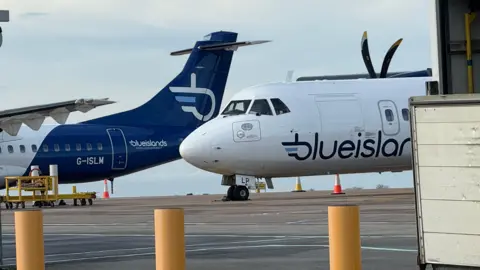 Two Blue Islands aircrafts on the tarmac at Guernsey Airport. Clouded blue skies can be seen behind.
