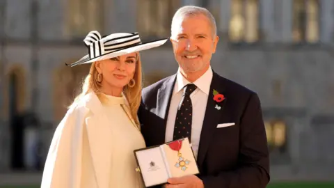 Reuters Karen Souness is wearing a white hat with black stripes, along with a white jacket and dress. She has shoulder length blonde hair. Graeme Souness wearing a dark suit and is holding his CBE in his hand. Both are smiling at the camera.