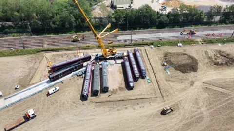 John Fairhall/BBC Train carriages on a piece of land next to a railway, with a couple being moved on to a make shift road