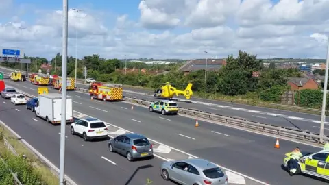 Scene of a crash where the A27 meets the M27. Both carriageways are closed and there is a line of emergency vehicles and a yellow air ambulance helicopter on the road. In the foreground is a queue of cars on the slip road