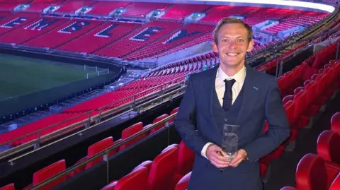 supplied A proud young man in a smart navy suit stands smiling and holding up an award surrounded by many rows of red seats in a huge football stadium