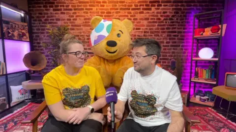 BBC Caroline Densley and Michael Chequer sat on chairs in a room, both wearing Children in Need shirts, and looking at eachother. Pudsey is stood behind them. Michael Chequer is holding a microphone. The wall behind them is brick. There are shelves behind them. The carpet on the floor is red.