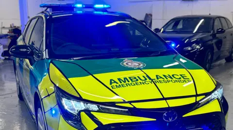 EEAST An ambulance, emergency response vehicle, in yellow and orange in a room, with two vehicles behind it. 