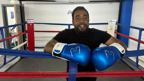 Jamal Choudhury - a man with black hair and short black stubble - is wearing bright blue boxing gloves and resting them over the side of a boxing ring. He is smiling into the camera and wearing a black top.