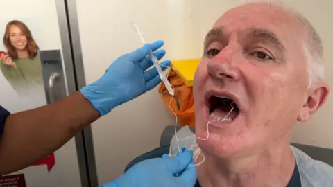 Ian Stewart in a medical apron sat in a chair at the mobile testing van. His mouth is open wide and there is a white string coming out of it, attached to a thin transparent stick being held by a nurse. 