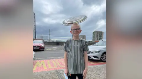 Smyth family A boy with blonde hair, wearing a grey stripy t-shirt, black shorts and glasses. He is smiling at the camera. He is standing in a car park.