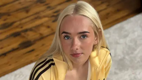 Miley Williams A blonde girl in a yellow tracksuit looks at the camera