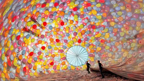 International Bristol Balloon Fiesta Two people stand inside an inflated envelope, that's the inflated part of the hot air balloon that sits on top of the basket. This one is very colourful with little tiny balloons designed in the style of the Disney film 'Up'.