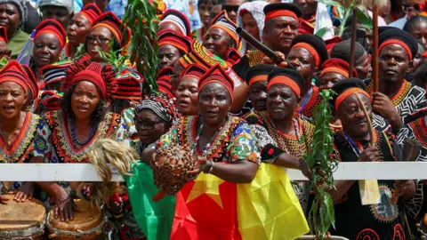 Reuters A crowd in Cameroon outside the cathedral in Bamenda dressed in colourful traditional outside and some with drums and calabash instruments.