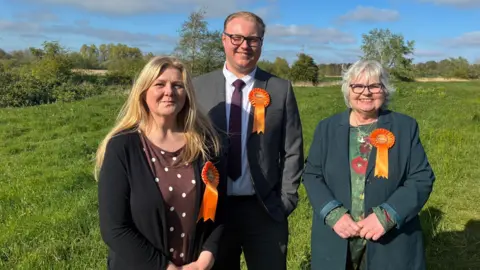 Dawn Merryweather, Jack Hilton, and Angela Greenfield standing in a field. They each wear a rosette. Dawn has long blonde hair and is wearing a long black cardigan over a brown top with white spots. Jack is wearing a grey suit with a white shirt and burgundy tie. He has black-rimmed glasses on and light blonde hair swept back. Angela is wearing a green jacket and floral green top and has white hair.