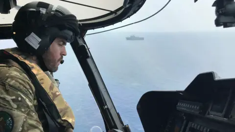 Steve Irwin Steve Irwin in a helicopter above the sea wearing camouflage uniform and a helmet, with a military-type ship in the far background