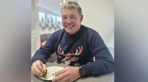 A man smiles at the camera, wearing a Christmas jumper while eating a mince pie at a kitchen table 