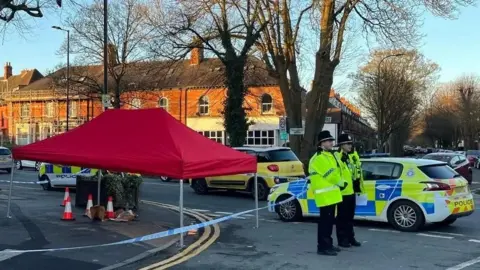 Police officers wearing hi-vis jackets stand in front of a cordon and red tent. Marked cars are parked in a road. Victorian buildings and tall, bare trees can be seen under a blue sky.