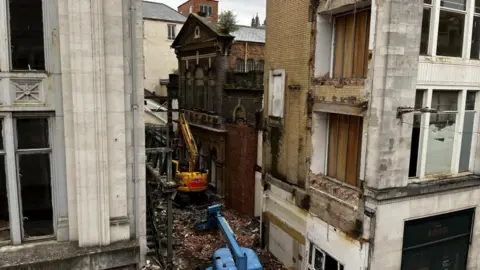The outside of a chapel building with rubble and diggers in front of it and other buildings either side.