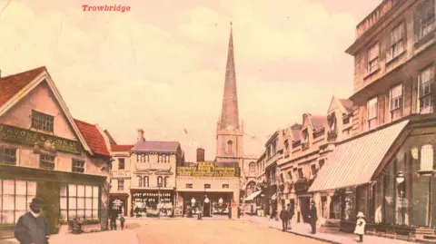 Wiltshire and Swindon History Centre P54130 Old postcard of a high street with one building in the middle which says 'KNEES' in yellow writing. A church spire is in the background