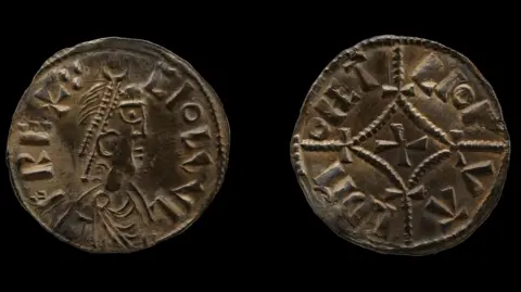 British Museum The photo shows both sides of a small coin, made of silver. It has the profile image of a king on one side and a cross on the other. Both sides include lettering.
