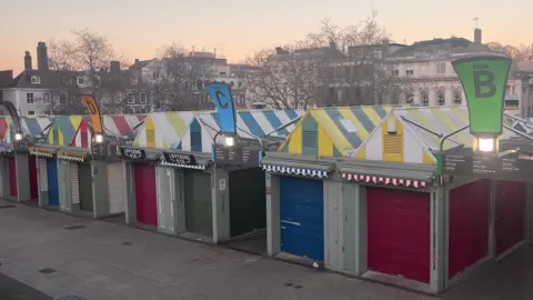 Neve Gordon-Farleigh/BBC Norwich markets. Rows of market stalls with coloured metal shutters and striped colourful roofs. 