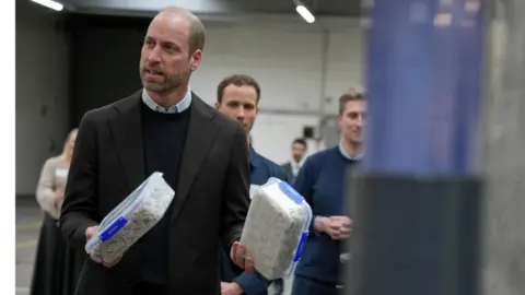 PA Media Prince William holds two Tuppaware boxes containing a cotton-wool like substance. He wears a black suit and black sweater. Other people are visible in the background. 