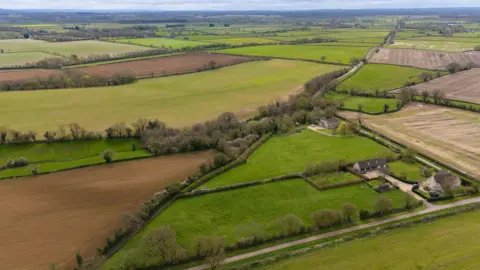 An aerial view of green land. Acres of land can be seen lined with trees and there is a road and a couple of houses also visible.