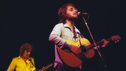 Getty Images Gerry Rafferty has a beard and is wearing glasses as he plays guitar and sings on stage 