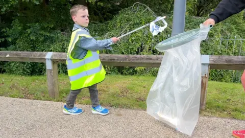 a young boy with a litter picker drops rubbish into a white bin bag. The boy has blue trainers, a denin shirt and a green hi vis top