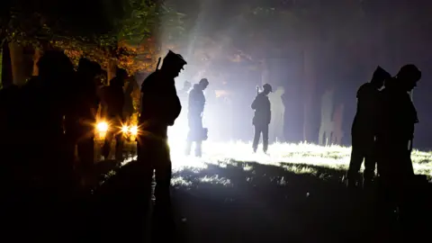 David Humphries A large number of silhouettes of servicemen in a garden, lit at night time. The silhouettes are black.