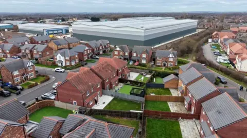 An aerial image of a large warehouse surrounded by houses, showing gardens and roads