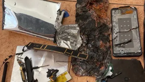Supplied A badly charred mobile phone, which is next to another device that has suffered significant damage and appears to have been dismantled. The items are laid out on an orange tile.