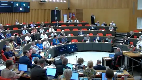 Cornwall Council The image shows a number of councillors sitting in a round, many with their laptops open, during a formal council meeting.