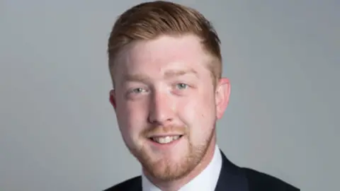 Corporate headshot of a man with reddish-brown hair and a trimmed beard and moustache smiling at the camera. A white short collar and black jacket are just visible at the bottom of the shot.