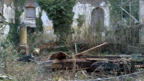 Whitfield Tabernacle Trust An old derelict building is pictured with rubble including overgrown trees, metal fences and a pond. 