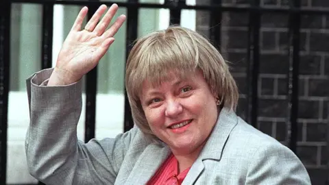 Getty Images Close-up of Mo Mowlam smiling and waving as she enters 10 Downing Street. She has fair hair with a fringe and is wearing a light grey suit jacket and red top.